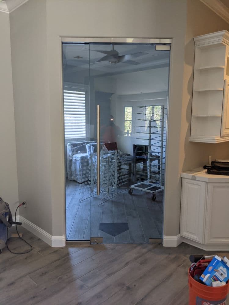 Frameless glass door with floor-mounted pivot hardware and a tall vertical pull, opening from a hallway into a back room