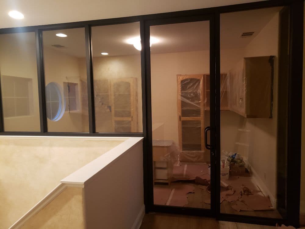 Black-framed tempered glass partition wall and door closing off a room mid-renovation, with fixed glass panels above the stair rail